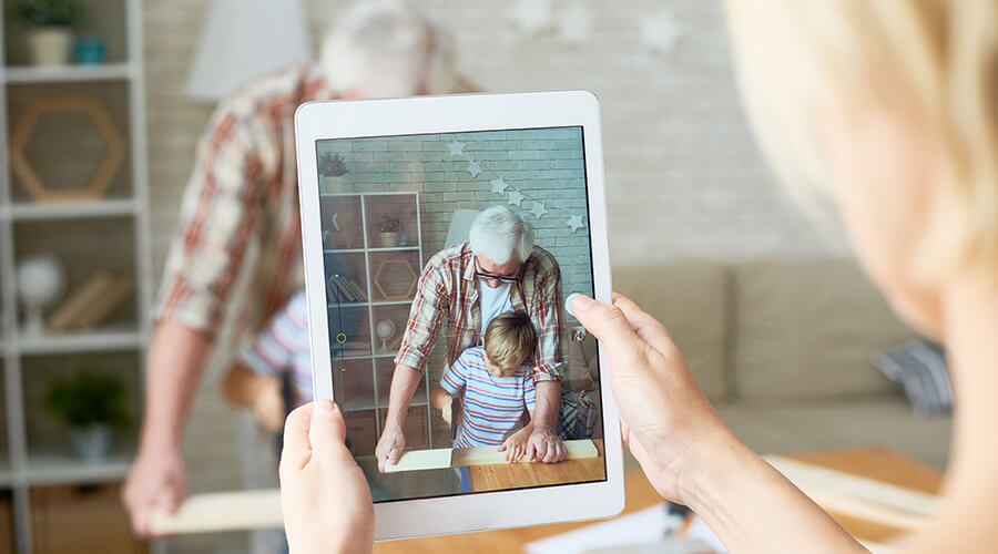 Woman holding tablet taking photo of grandfather and grandson doing crafts.